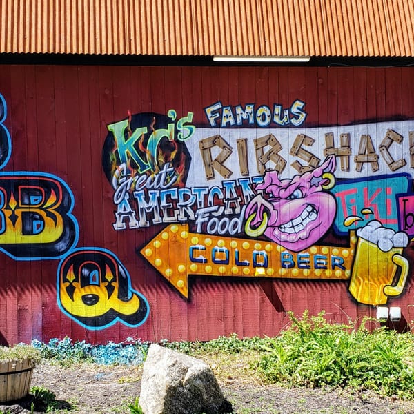 Colorful graffiti on a red wooden wall advertises "KC's Famous Rib Shack" and "Cold Beer" with a pig mascot.