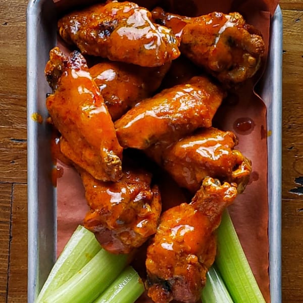 A metal tray of KC's Rib Shack BBQ chicken wings with celery sticks on a wooden table.