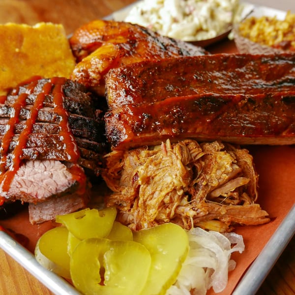A BBQ platter with ribs, brisket, pulled pork, cornbread, pickles, and coleslaw.